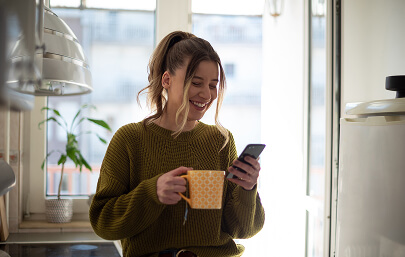 Woman holding a mug and smiling at her phone at home